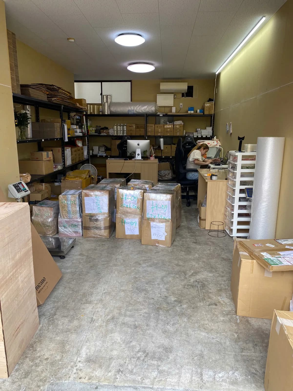 棚に梱包資材を備え、奥で出荷手配を進めるスタッフがいる倉庫内